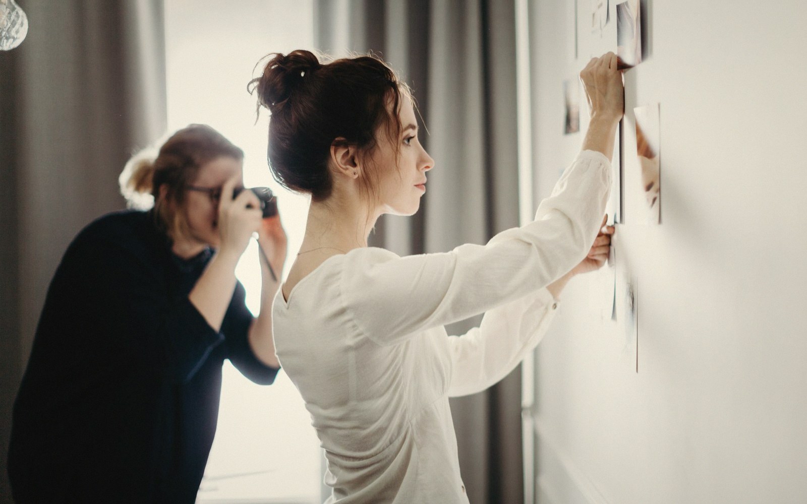A woman pins photos to a wall while another photographs her with a camera