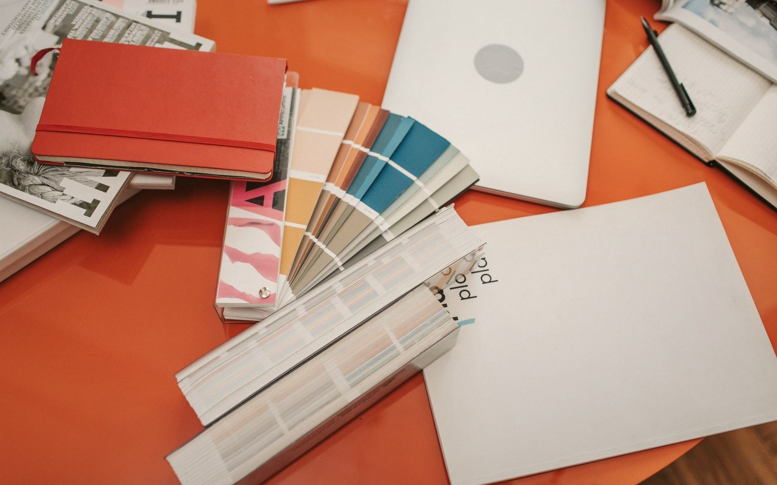 Paint swatch fans and notebooks spread across an orange desk beside a laptop