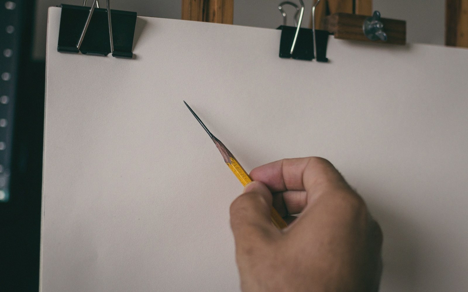 A hand holds a sharpened pencil above blank paper clipped to a drawing board
