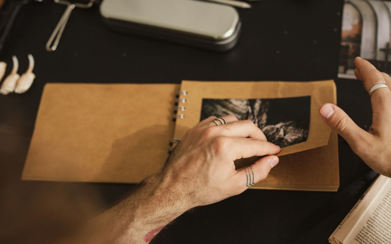 Hands with ring tattoos turn a page in a kraft-paper photo book on a dark table