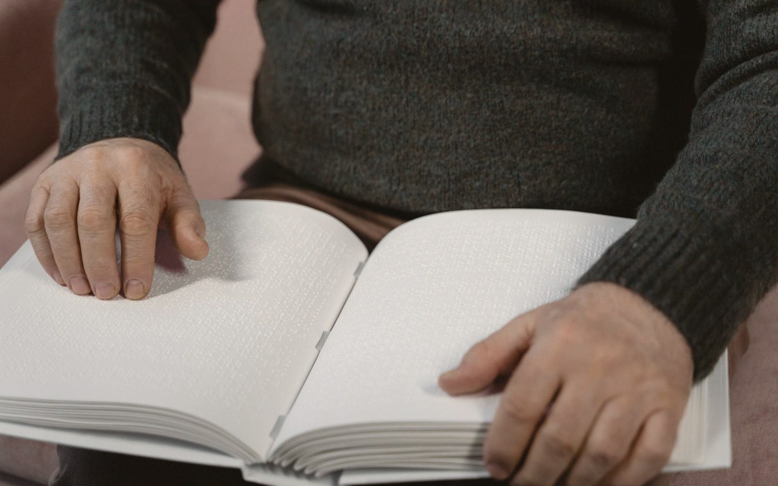 A person's fingers reading braille text in an open book on their lap