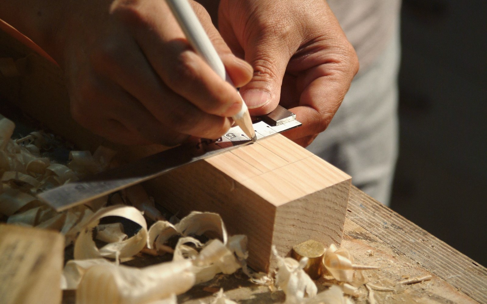 Hands using a pencil and square to mark measurements on a wooden block amid curled shavings